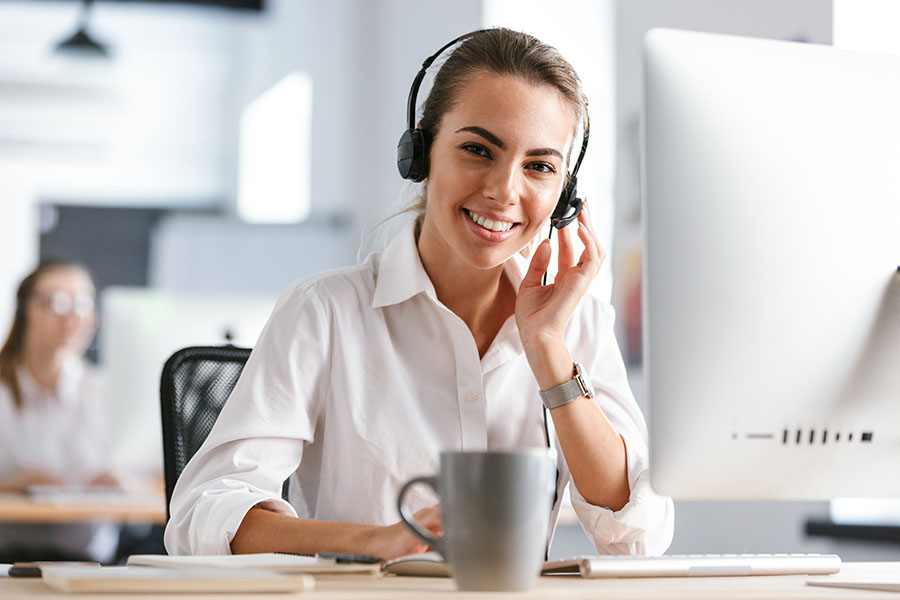 Smiling young woman wearing microphone headset dressed in shirt sitting at her workplace at the office, working on computer