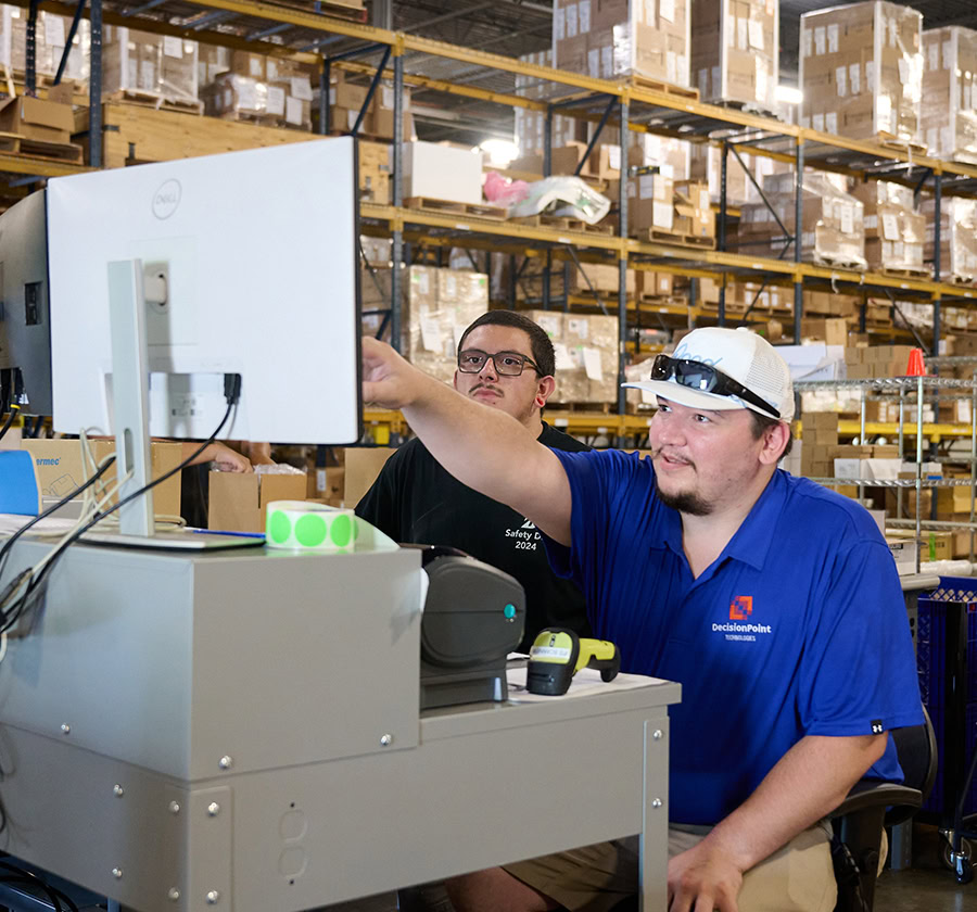 Two men in a warehouse review a computer screen, one in a blue DecisionPoint shirt.
