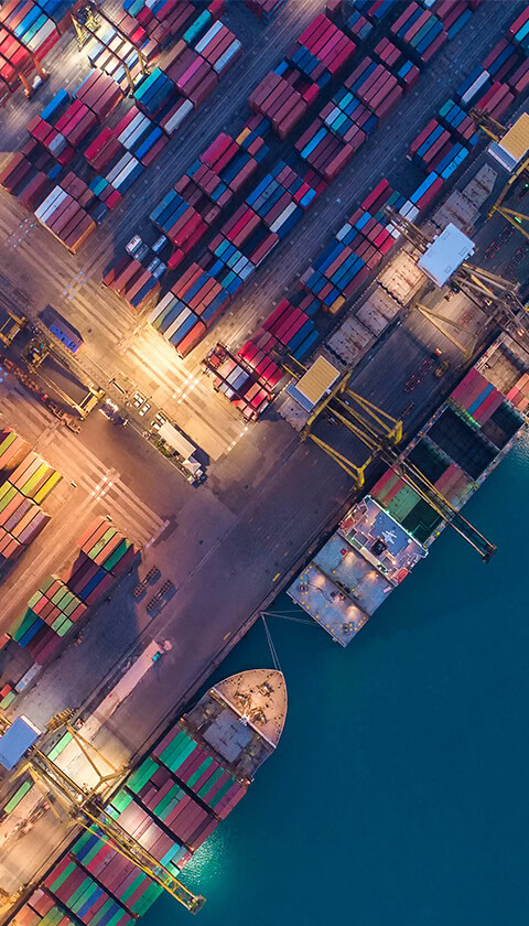 Overhead view of a shipping yard, showing docked ships and hundreds of stacked shipping containers.